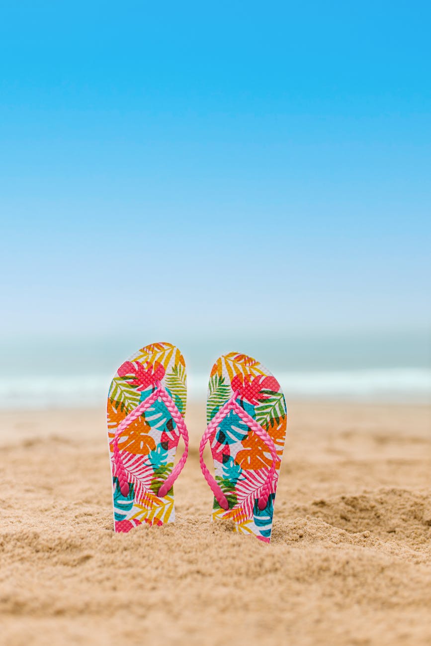 Vibrant flip flops stand in the sand against a clear blue sky, symbolizing a perfect summer beach day.