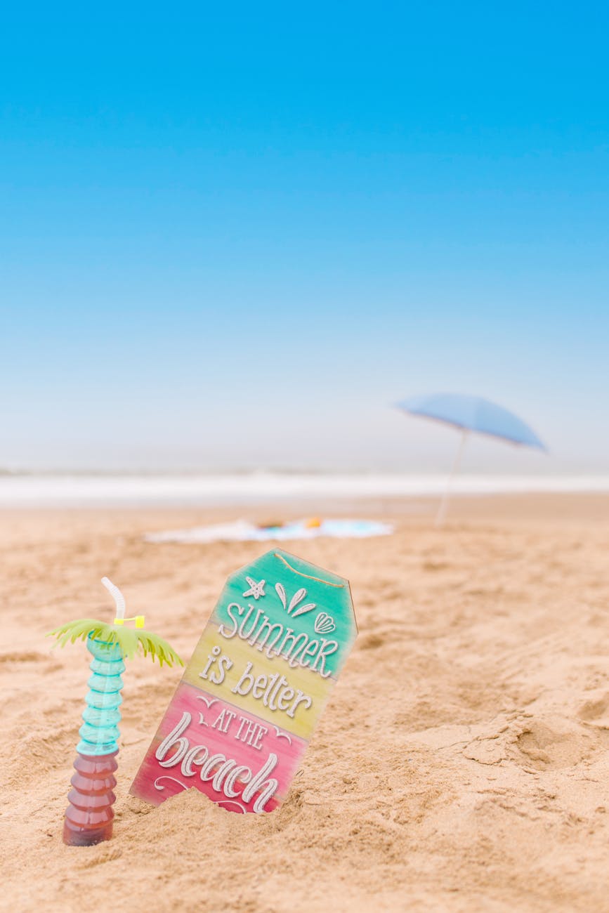 Vibrant beach sign and drink capture summer vibes on sandy beach with blue sky.