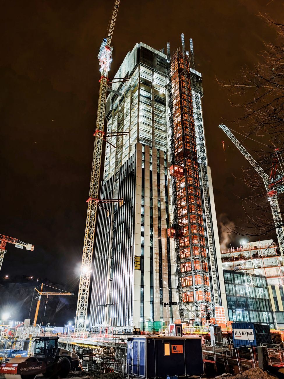 A modern skyscraper under construction at night in Lindholmen, Sweden, illuminated by cranes and lights.