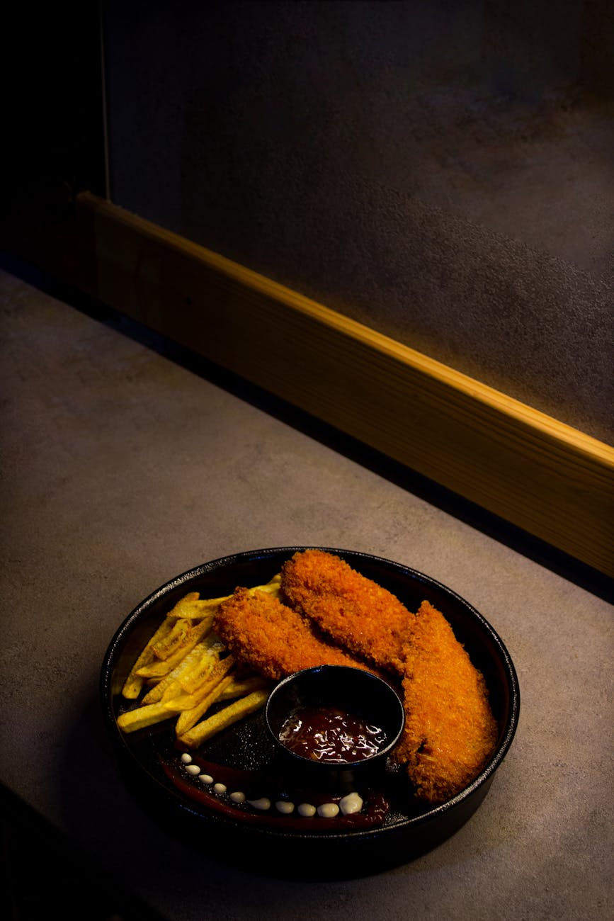 Appetizing plate of crispy fried chicken and fries with dip served on a dark background.