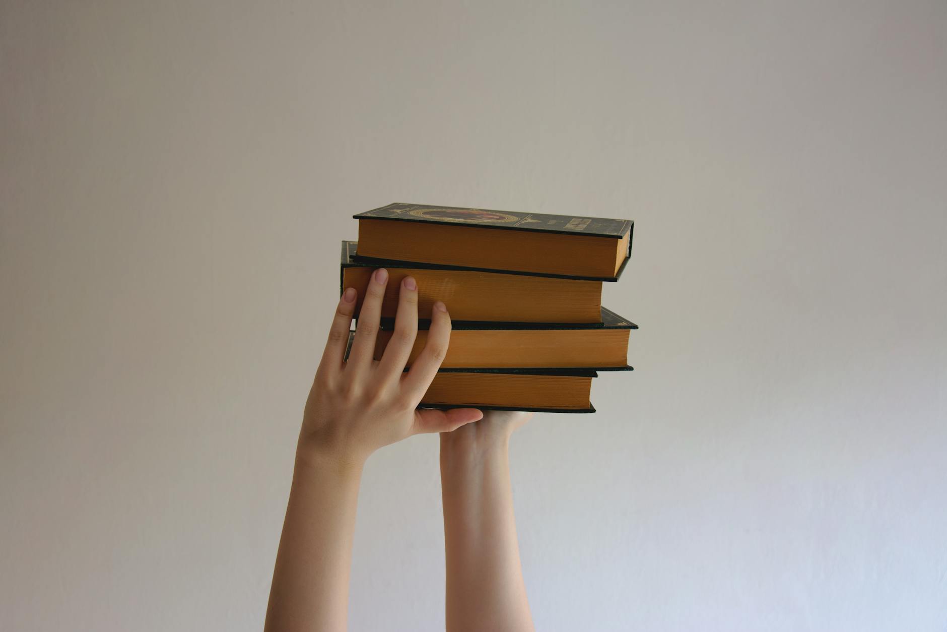 Hands reaching upwards holding a stack of hardbound books against a plain background.