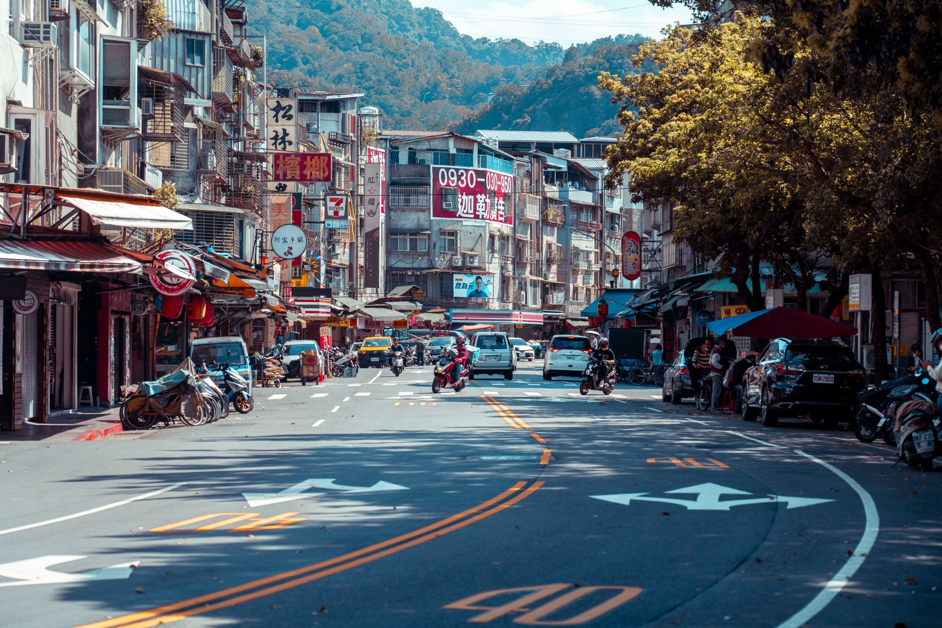 Vibrant urban street scene with shops, vehicles, and mountains in an Asian city.