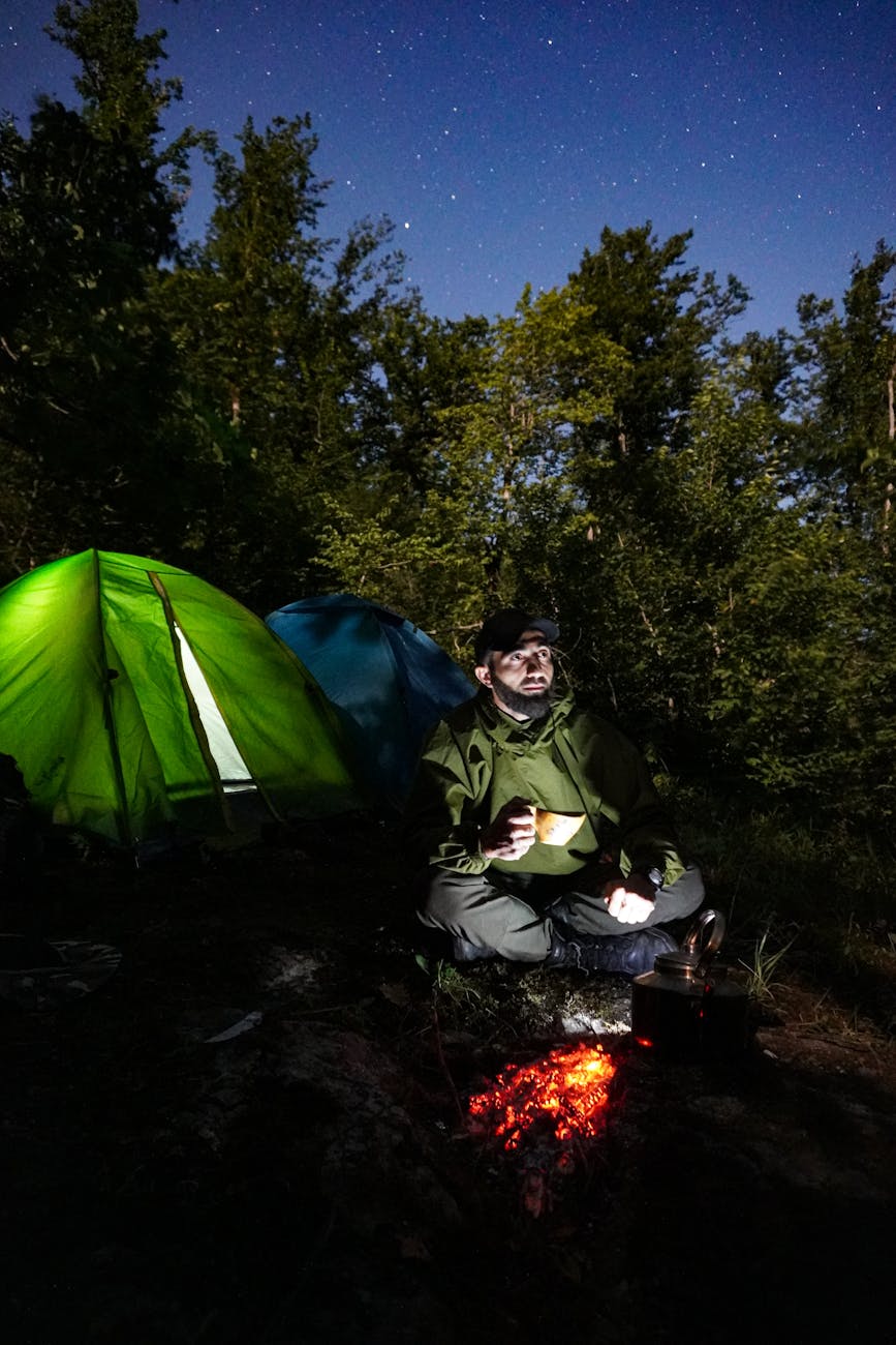 Outdoor camping scene with a man by the fire under starlit sky, surrounded by tents in a forest.