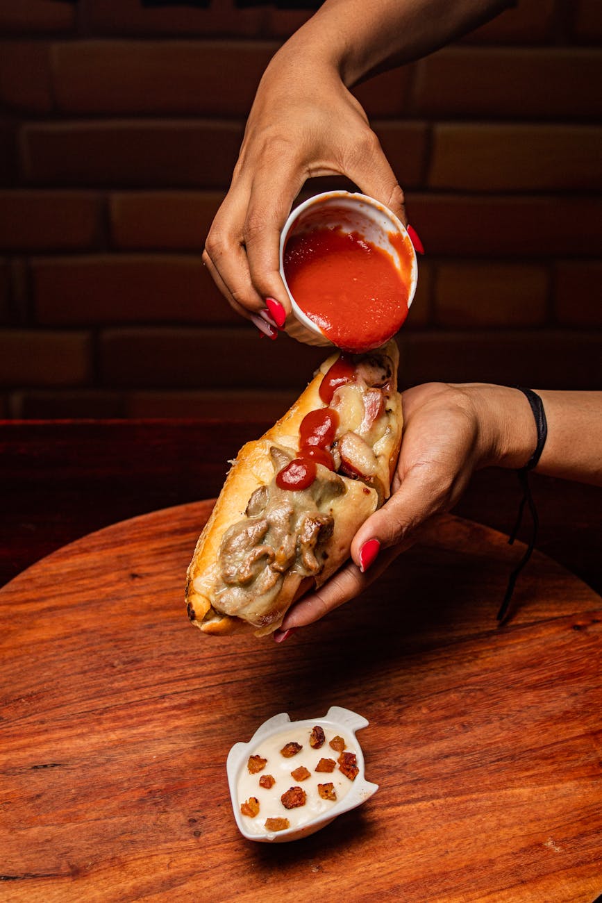 Close-up of a hot dog being topped with tomato sauce by hand over a wooden table.