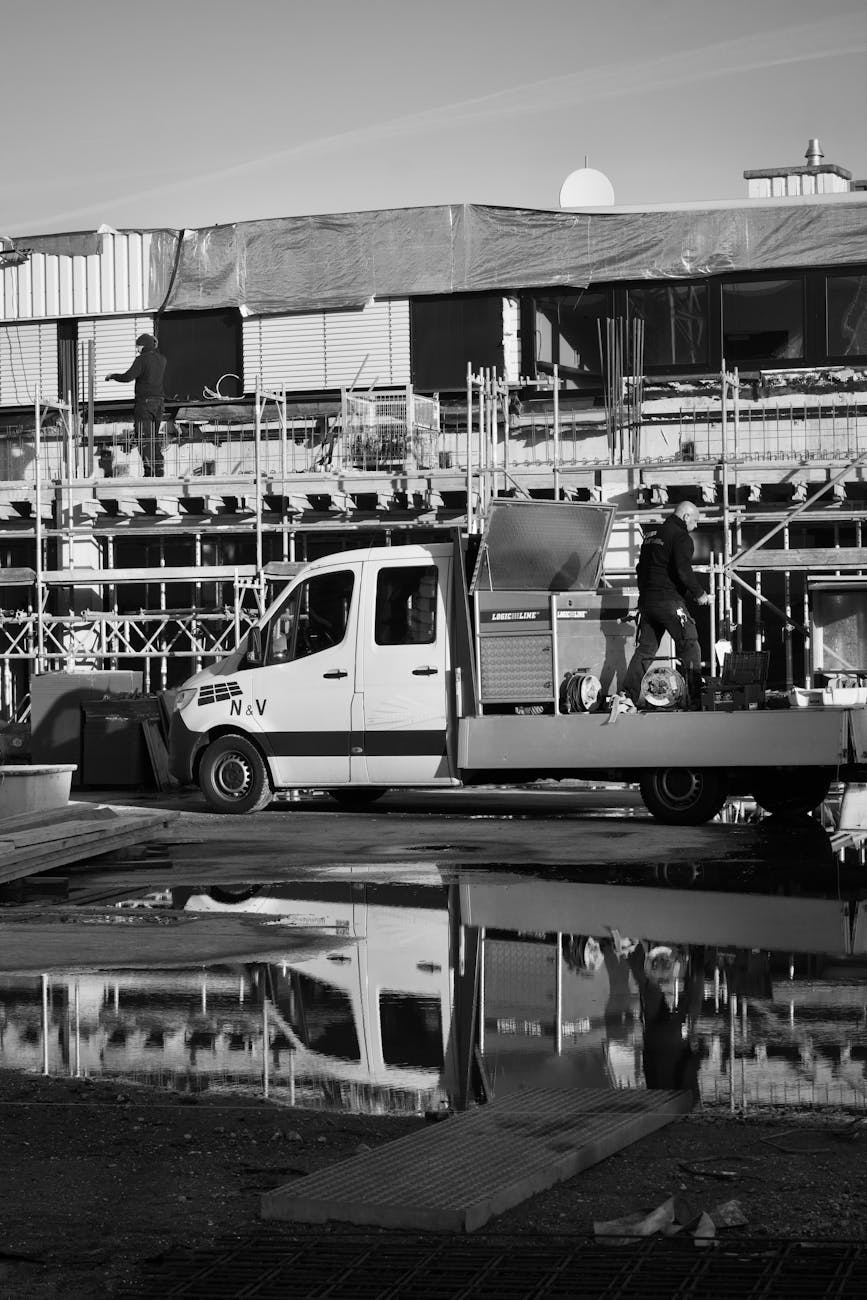 Monochrome image of a construction site with workers and vehicles reflecting in water.
