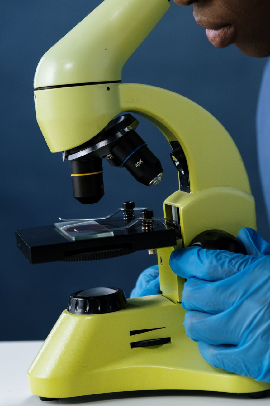 A close-up view of a microbiologist examining a sample under a microscope wearing blue gloves.