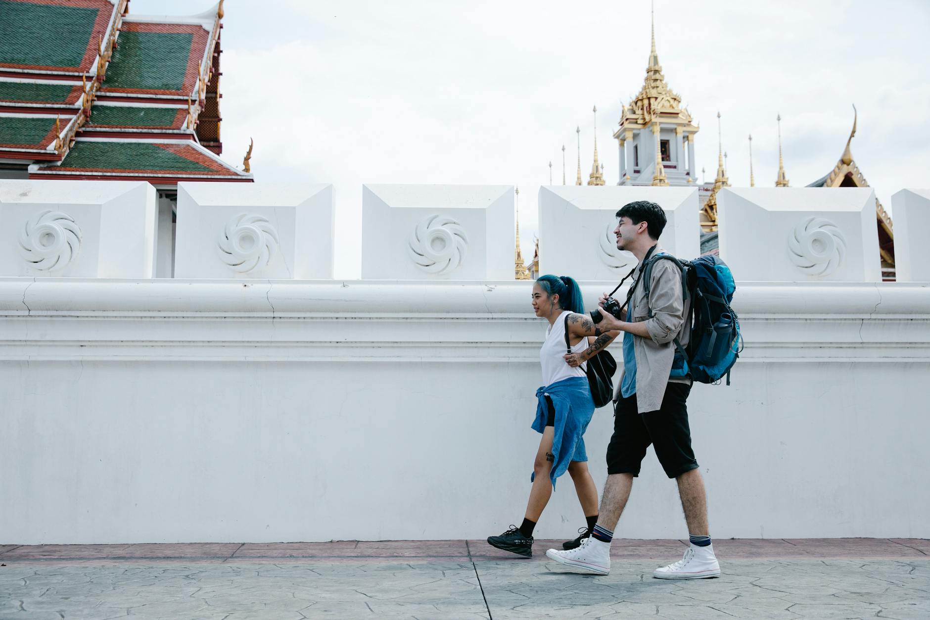 A couple with backpacks walks past stunning Thai temple architecture in an urban setting.