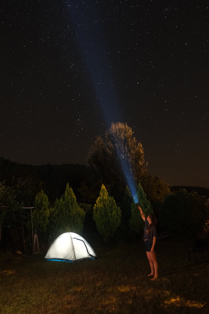 A woman shines a flashlight towards a starry sky beside a lit tent, capturing serene outdoor camping experience.