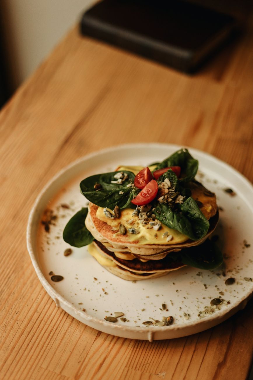 Stacked pancakes with cheese, spinach, cherry tomatoes, and seeds on a wooden table.