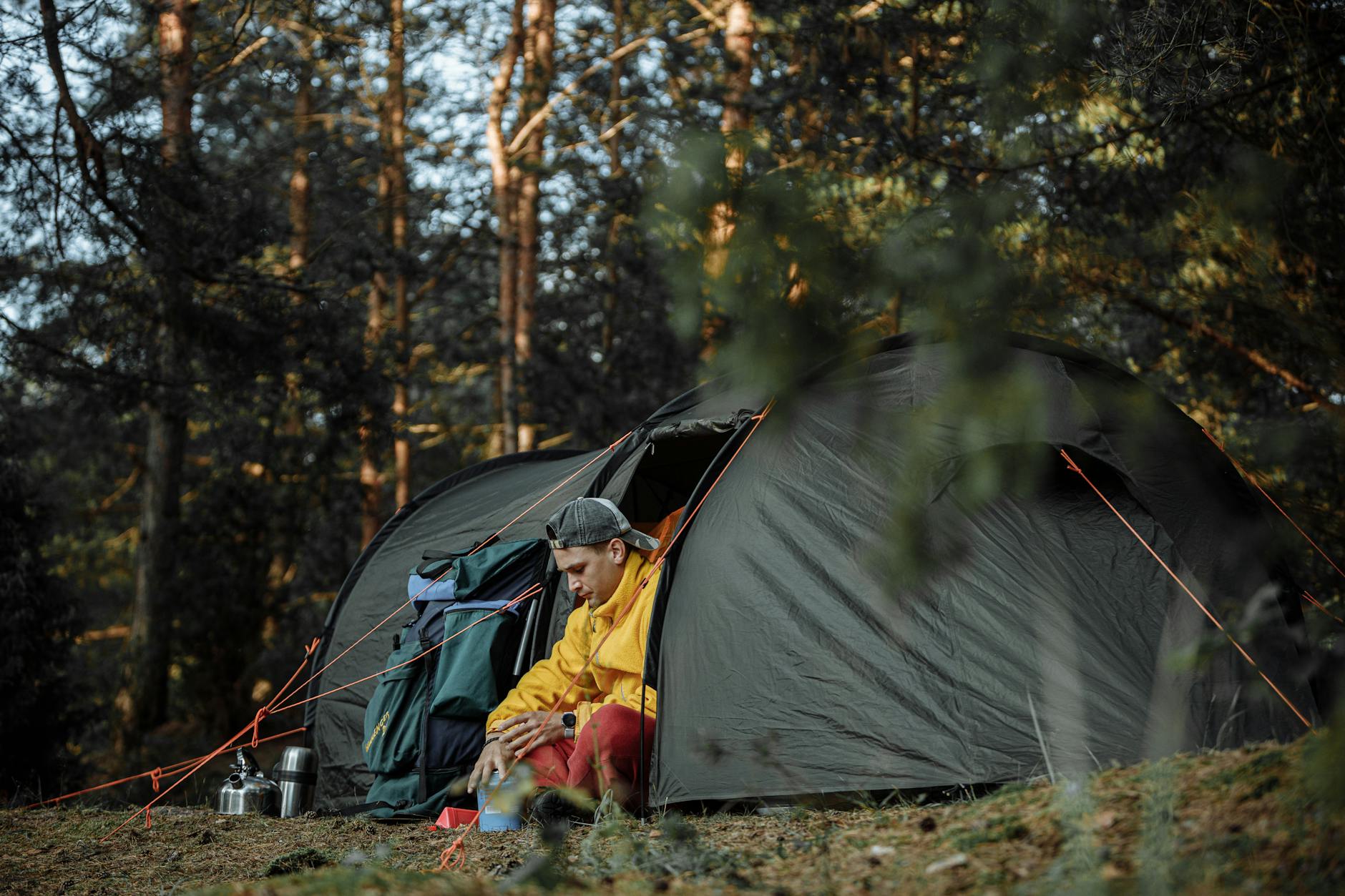 A man sitting by his tent in a forested campsite during the day.