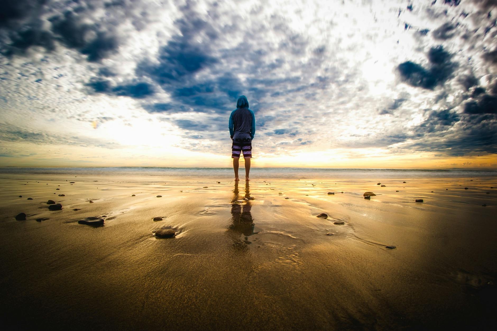 A lone figure stands on a tranquil beach during a vivid sunset, reflecting on the wet sand.