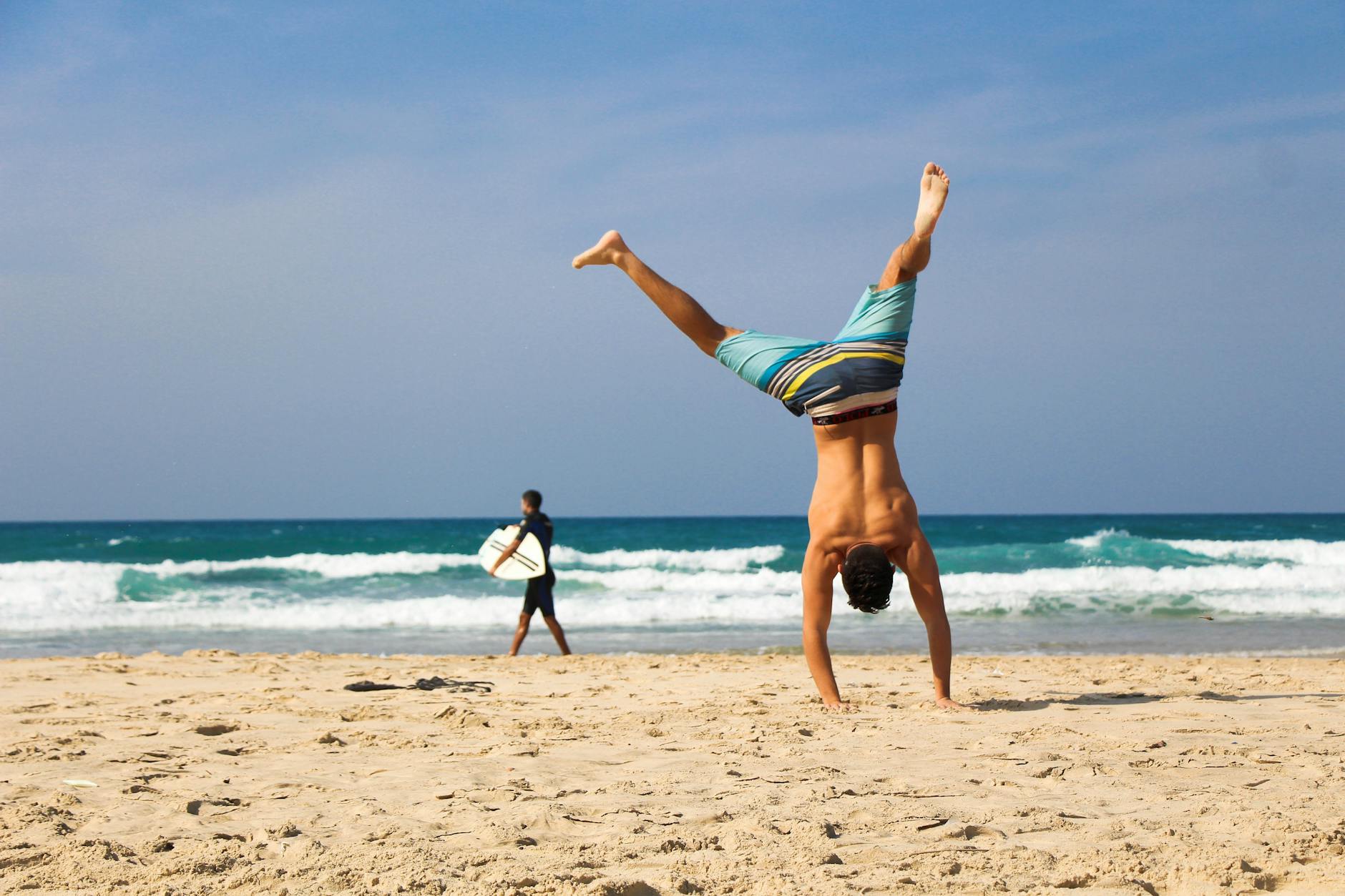 Man doing a handstand on a sunny beach with surfer in background, evoking a sense of freedom and joy.