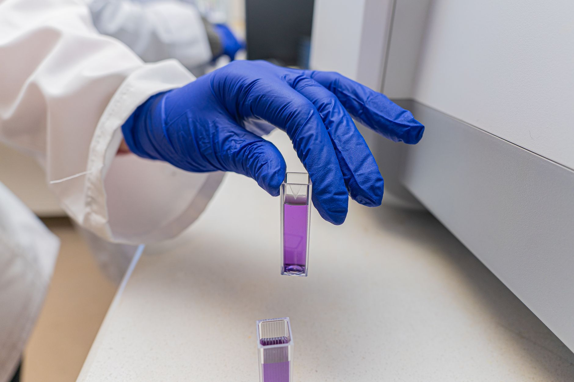 Close-up of a scientist's gloved hand handling a lab sample vial containing a purple liquid.