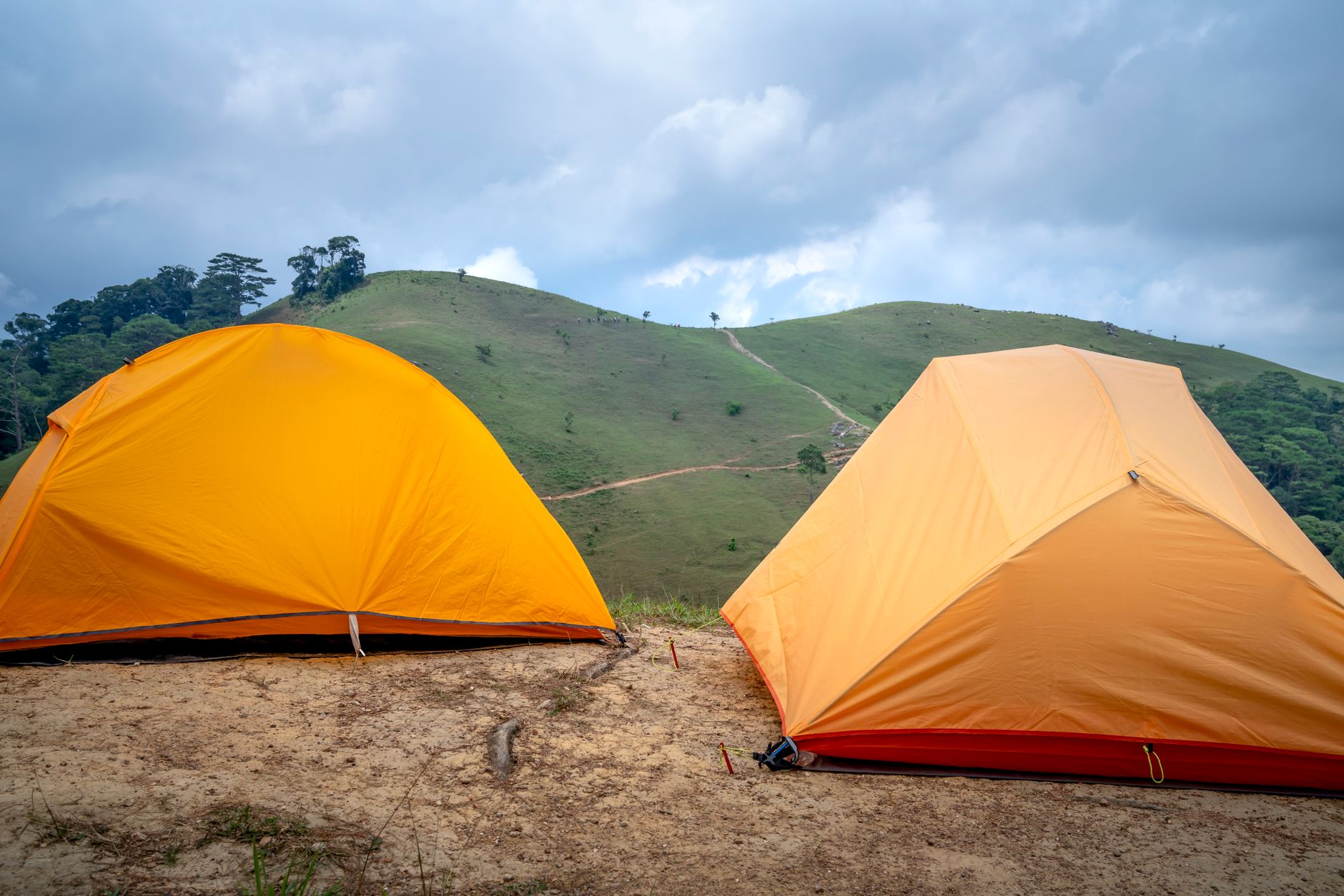 Colorful orange camping tents located on sandy ground near green mountain covered with grass against cloudy sky on summer day
