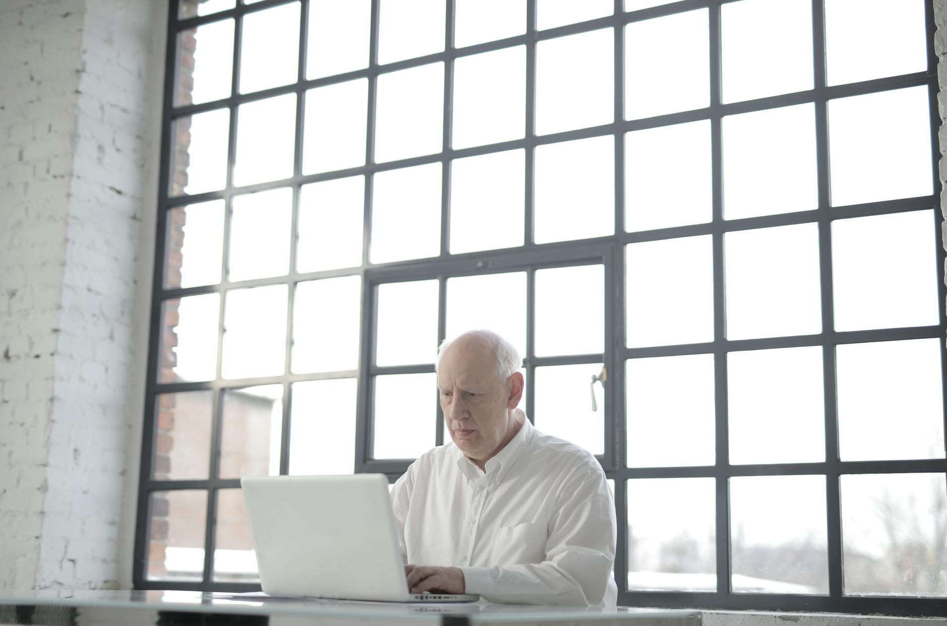 Focused senior man working on laptop in bright modern office with large windows.