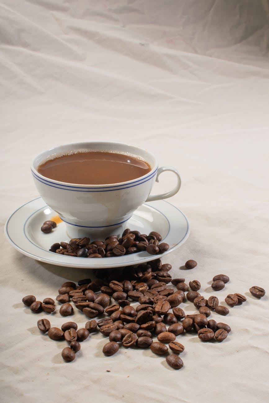 A porcelain cup filled with coffee surrounded by scattered coffee beans on linen.
