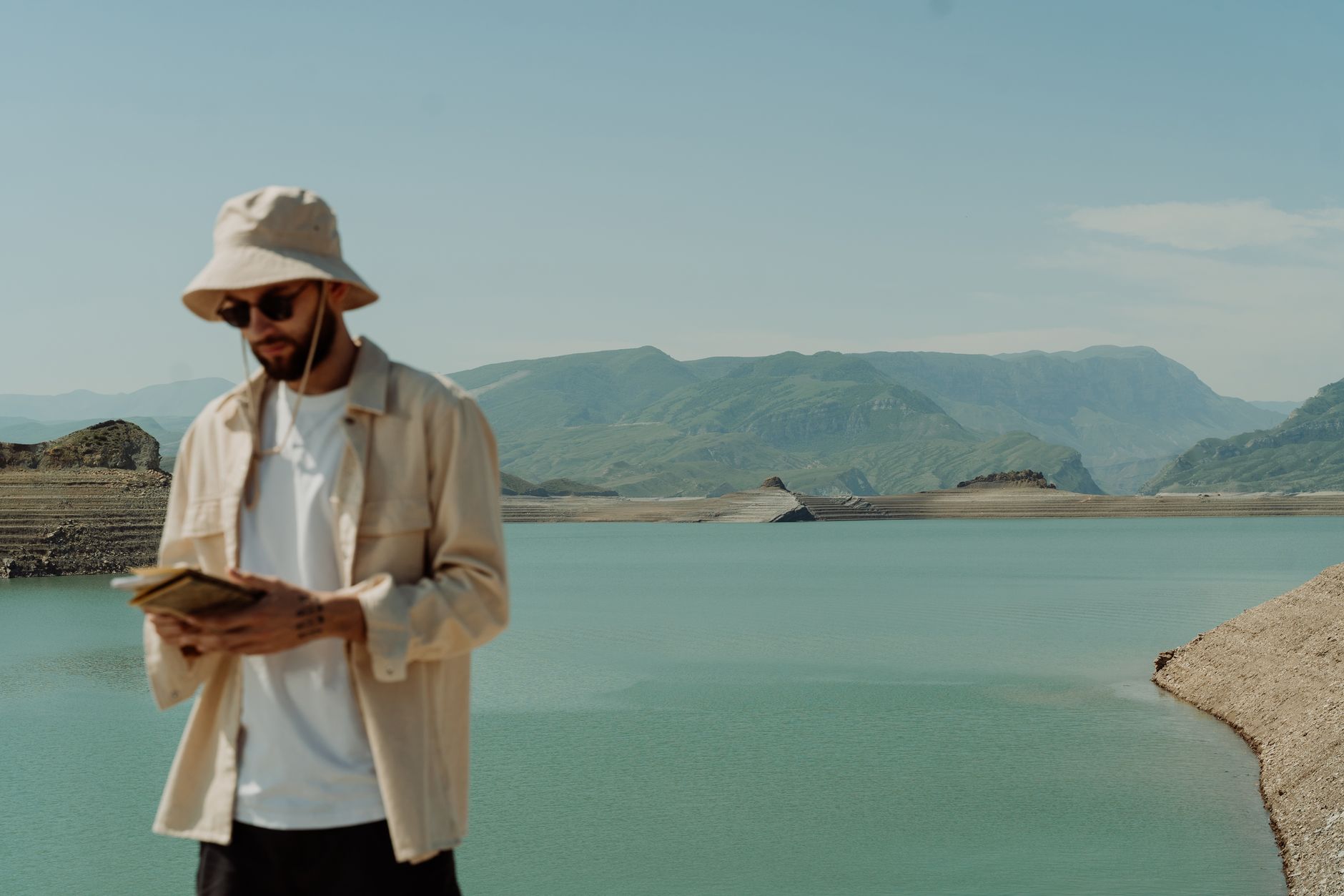 A man wearing a bucket hat and sunglasses looks at a book by a peaceful reservoir.