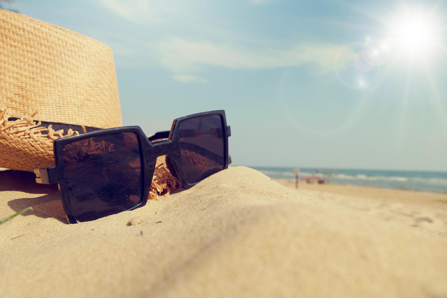 Close-up of sunglasses and straw hat on sandy beach under bright sunlight, perfect summer vibe.