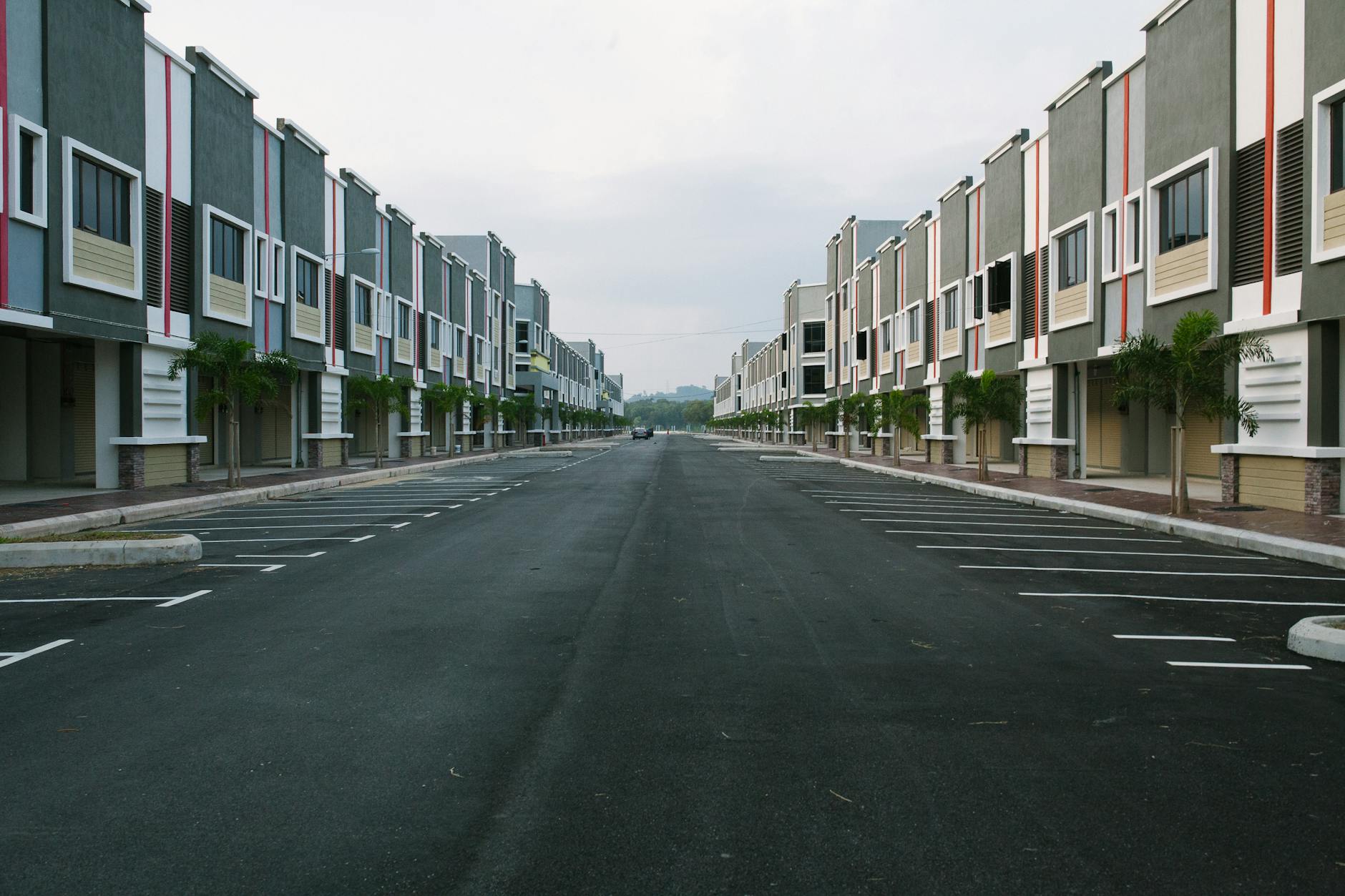 A quiet, empty urban street flanked by modern condominiums under a cloudy sky, with ample parking space.