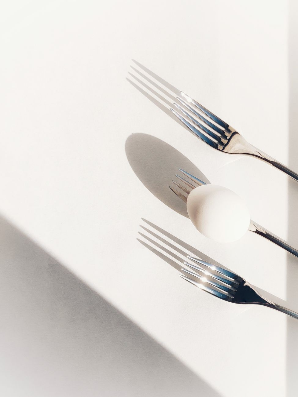 Artistic flatlay of an egg and three forks casting shadows on a white surface, top-down view.