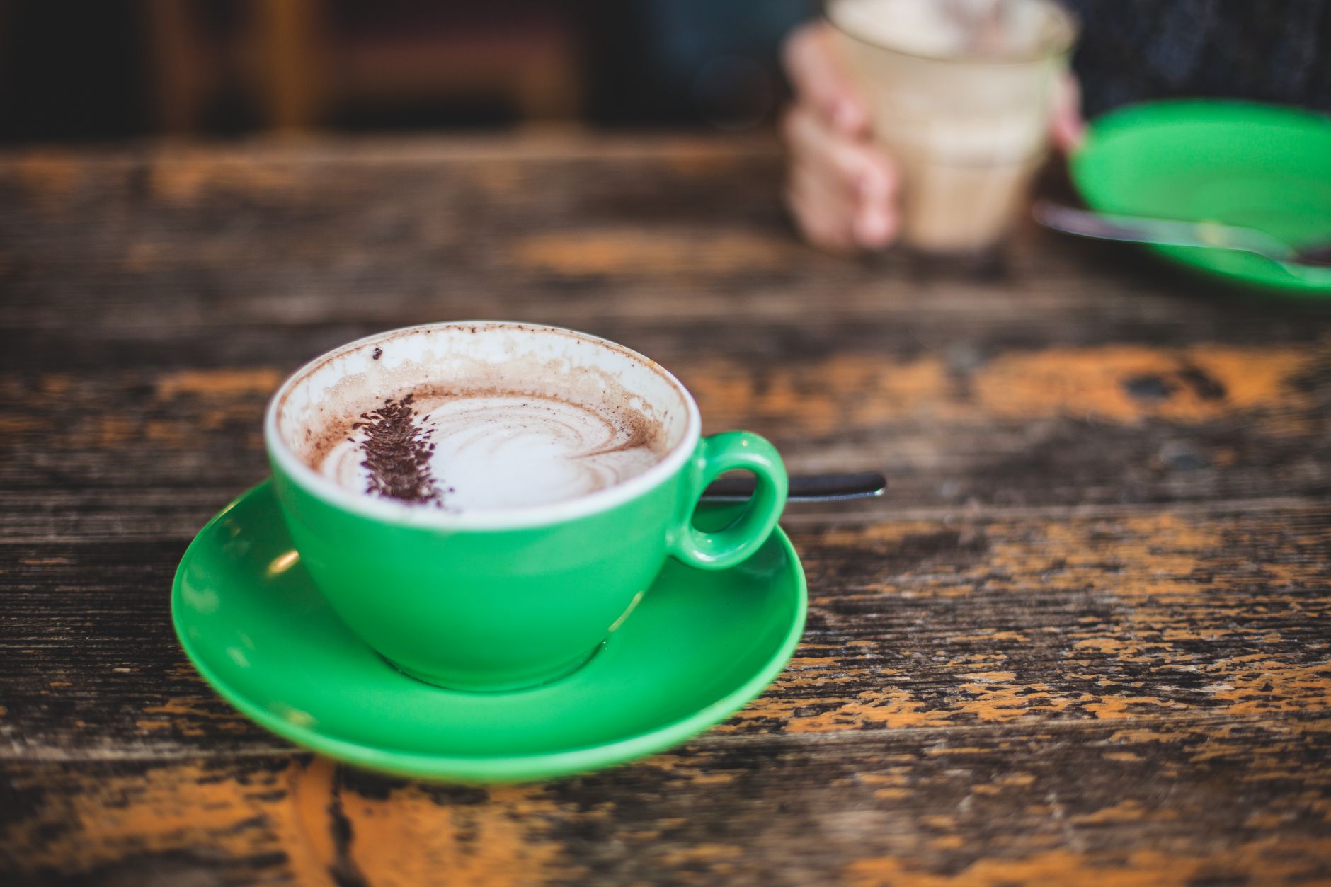 A vibrant green cup of cappuccino on a rustic wooden table with another drink in the background.