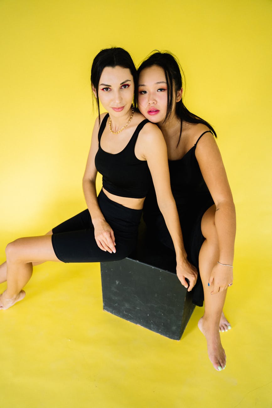 Two women in black outfits sitting on a black box against a vibrant yellow background.