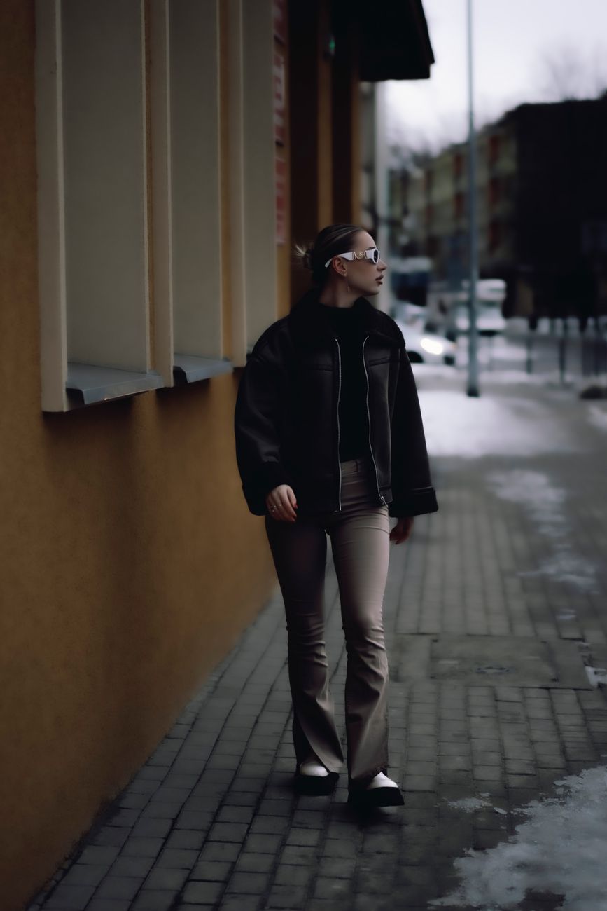 Fashionable woman strolls along snowy urban sidewalk in winter attire.
