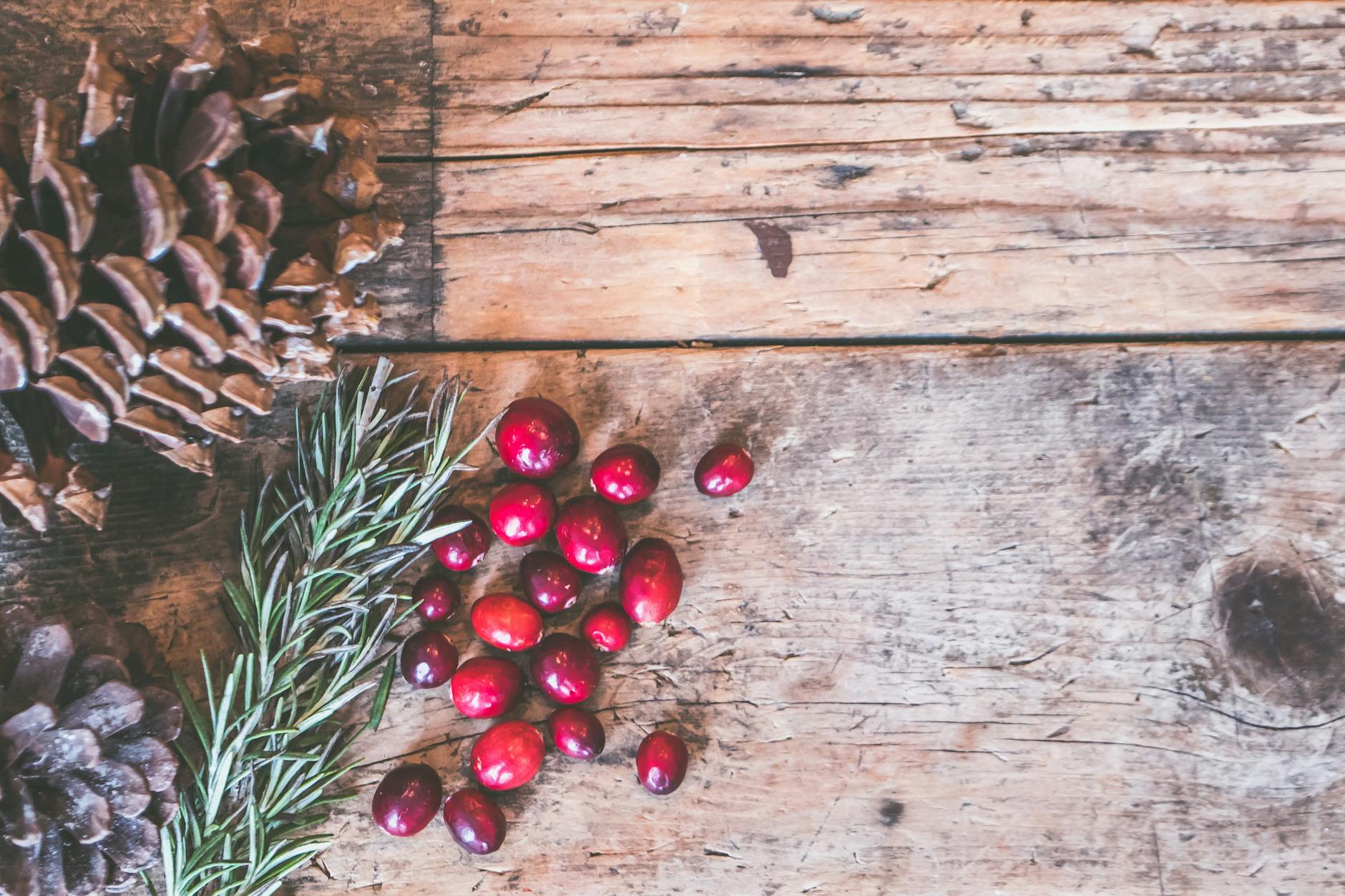 Top view of rustic holiday decor featuring pinecones, cranberries, and rosemary on a wooden surface.