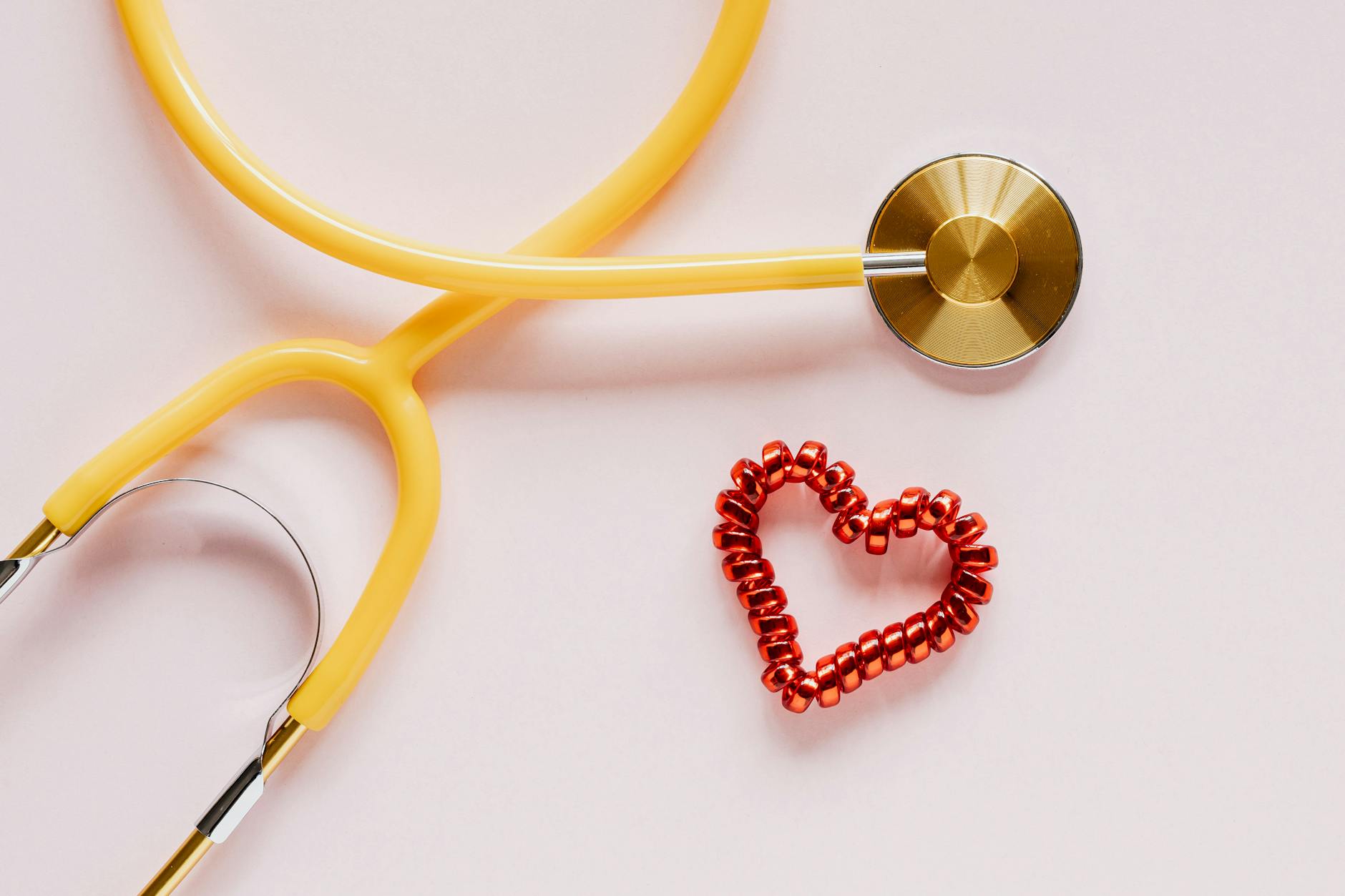 A yellow stethoscope and red heart decoration symbolize love and healthcare on soft pink.