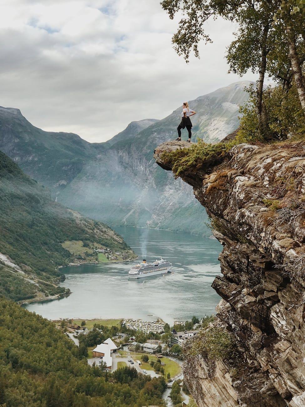 Person enjoys breathtaking view from a cliff above Geiranger Fjord, Norway, with clear skies and cruise ship below.
