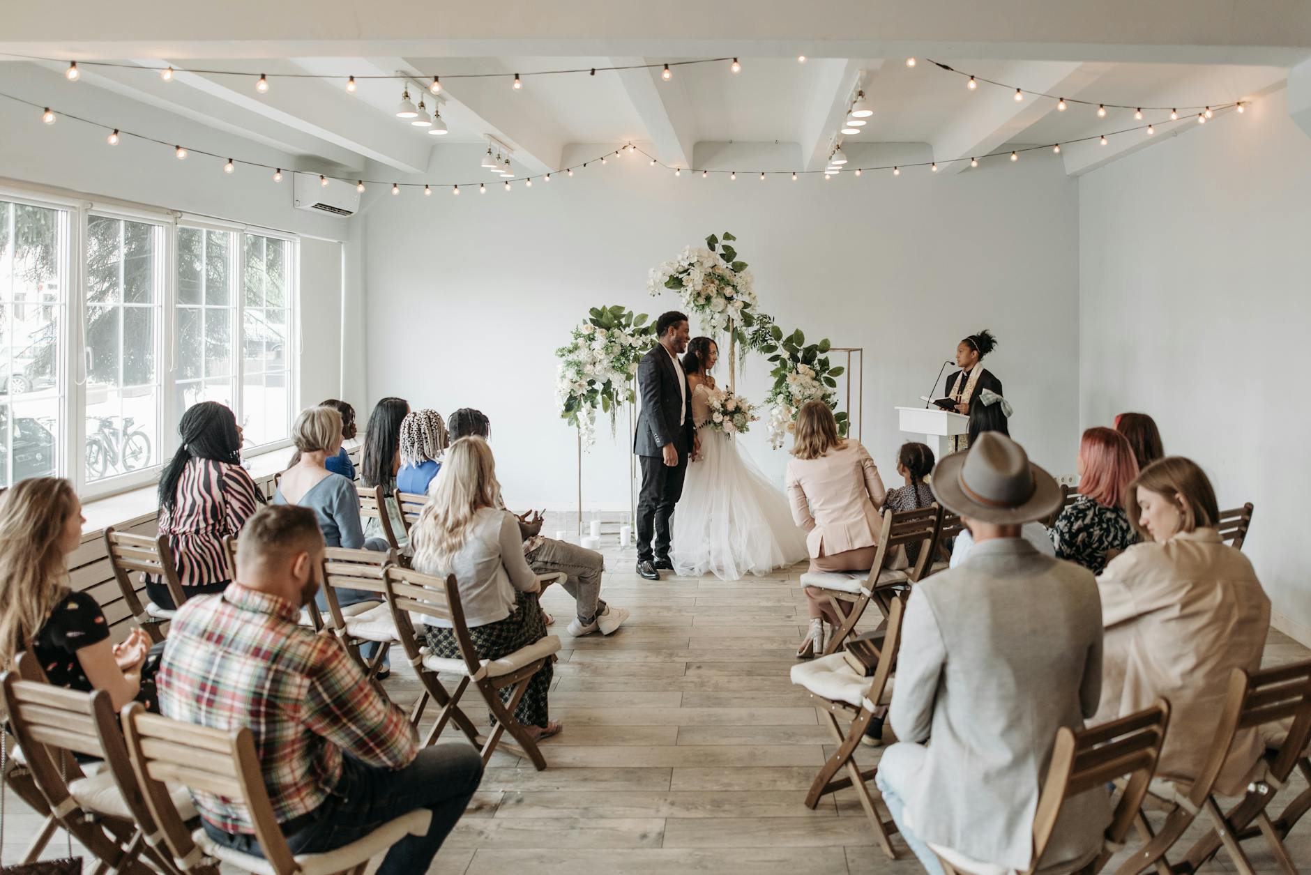 A small gathering witnesses a beautiful wedding ceremony indoors with wooden chairs and floral decor.
