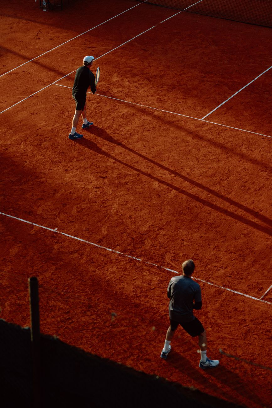 Two adults playing tennis on a red clay court during daytime. Shot from above, highlighting shadows.