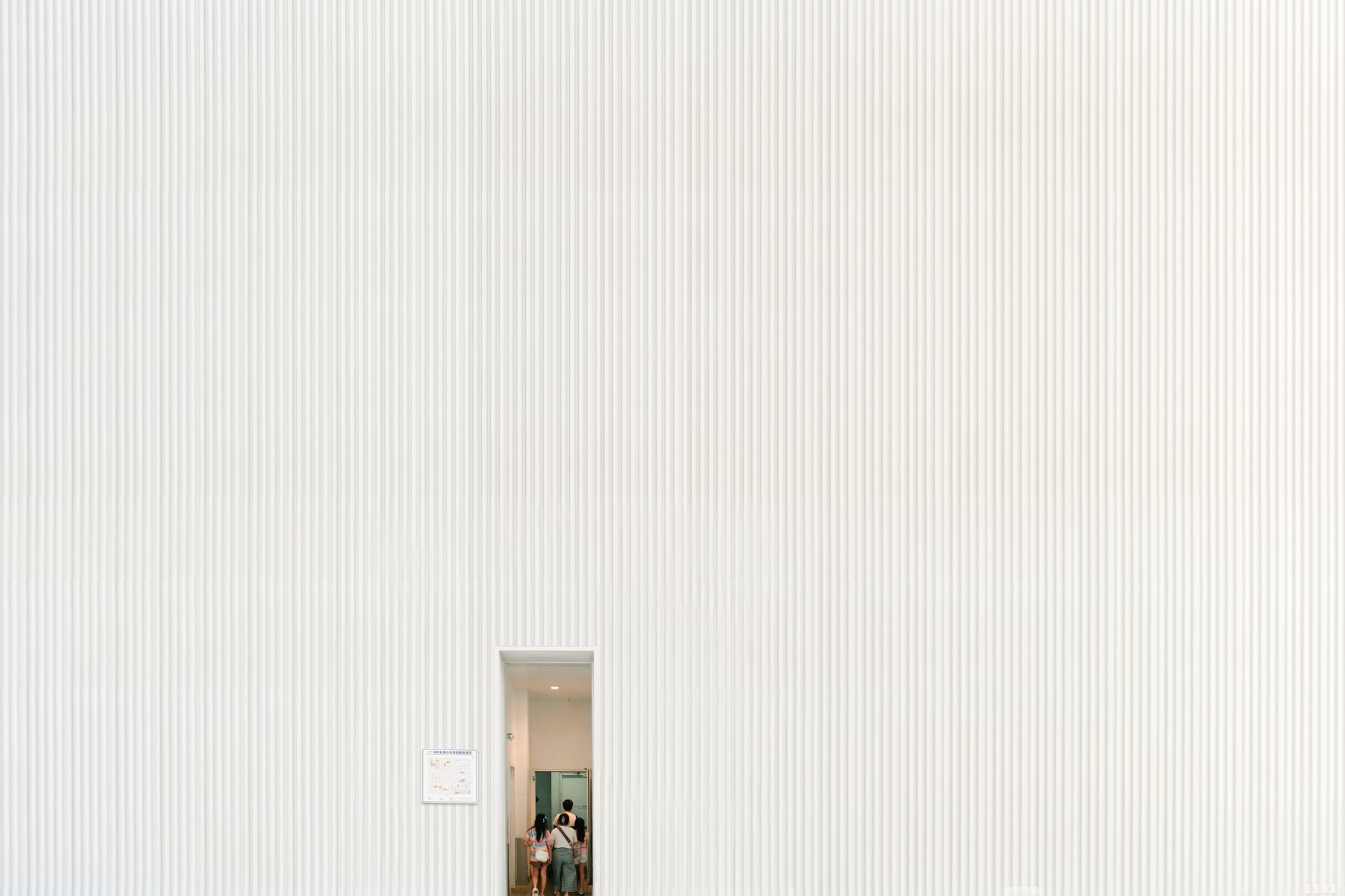 A large white textured wall with a small doorway entrance and visible people inside.