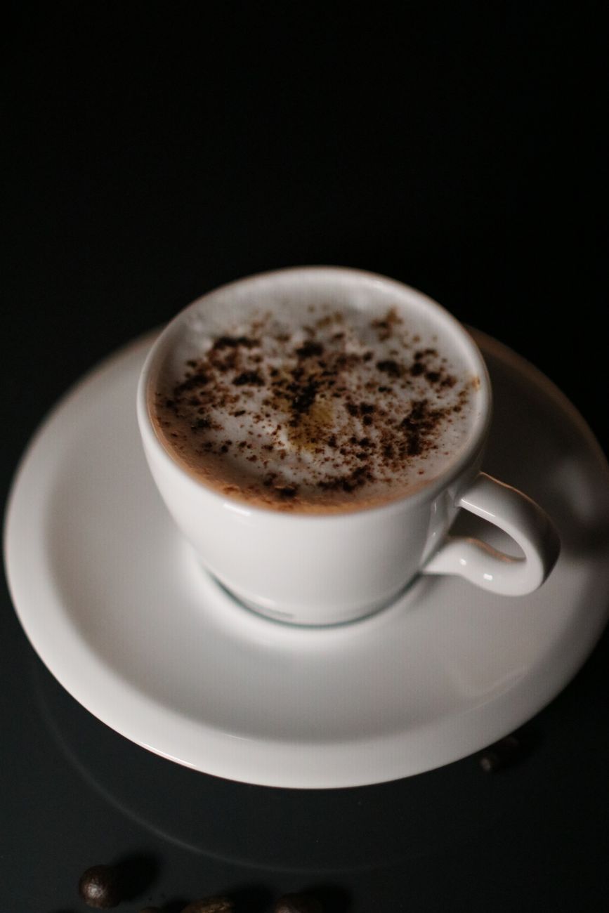 A ceramic coffee cup with frothy top dusted with cocoa powder, shot against a dark background.