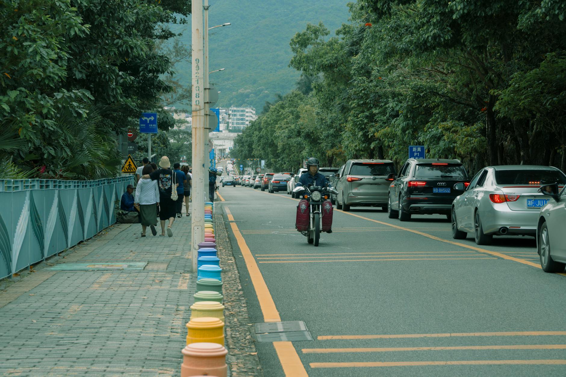 A busy city street with motorbike, cars, and walking pedestrians on a tree-lined road.