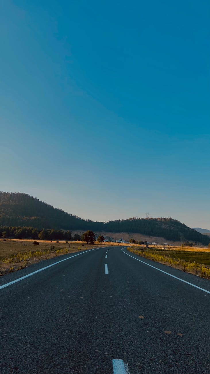 A scenic countryside highway stretches under a vast clear blue sky, surrounded by lush fields.
