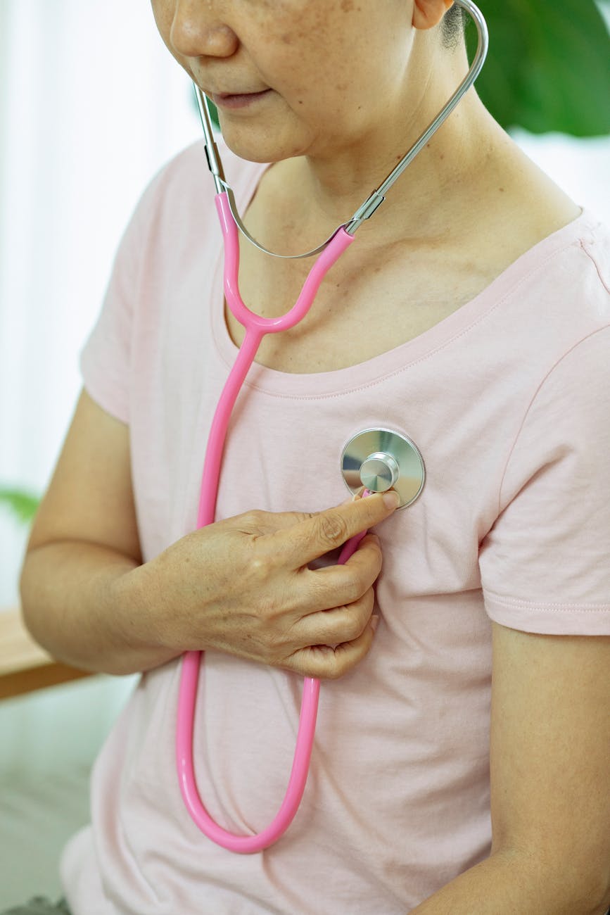 Crop unrecognizable serious middle age female in casual outfit using pink stethoscope on chest while sitting in bright house