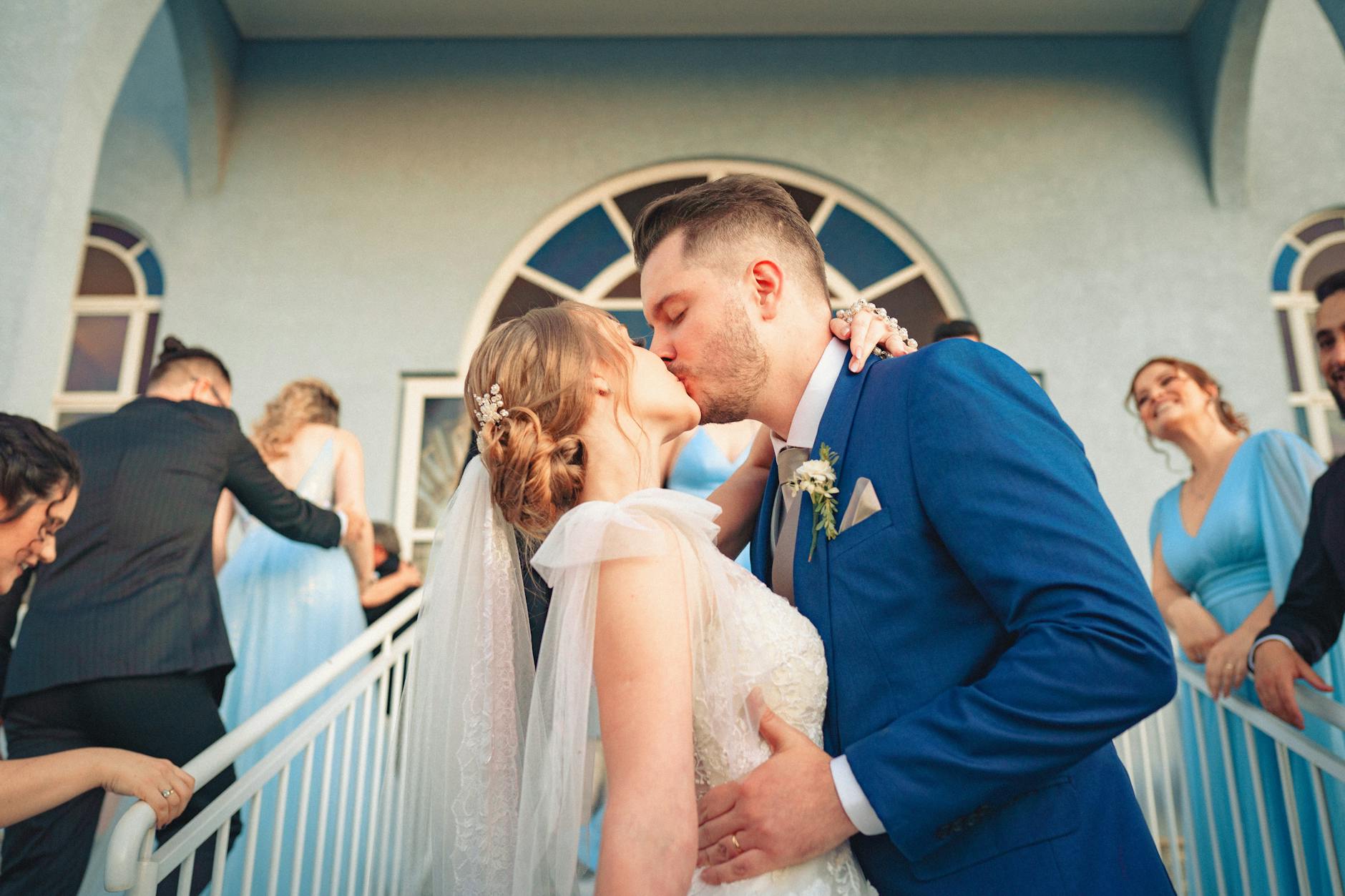A couple shares a romantic kiss at their wedding, surrounded by guests in formal attire.