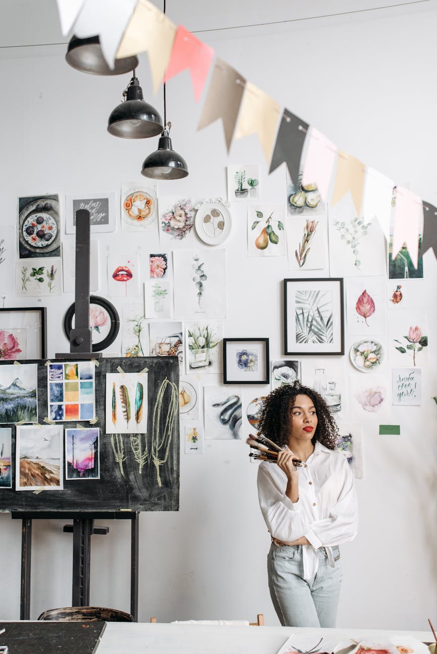 A woman art instructor in a workshop holding brushes, surrounded by art and drawings.