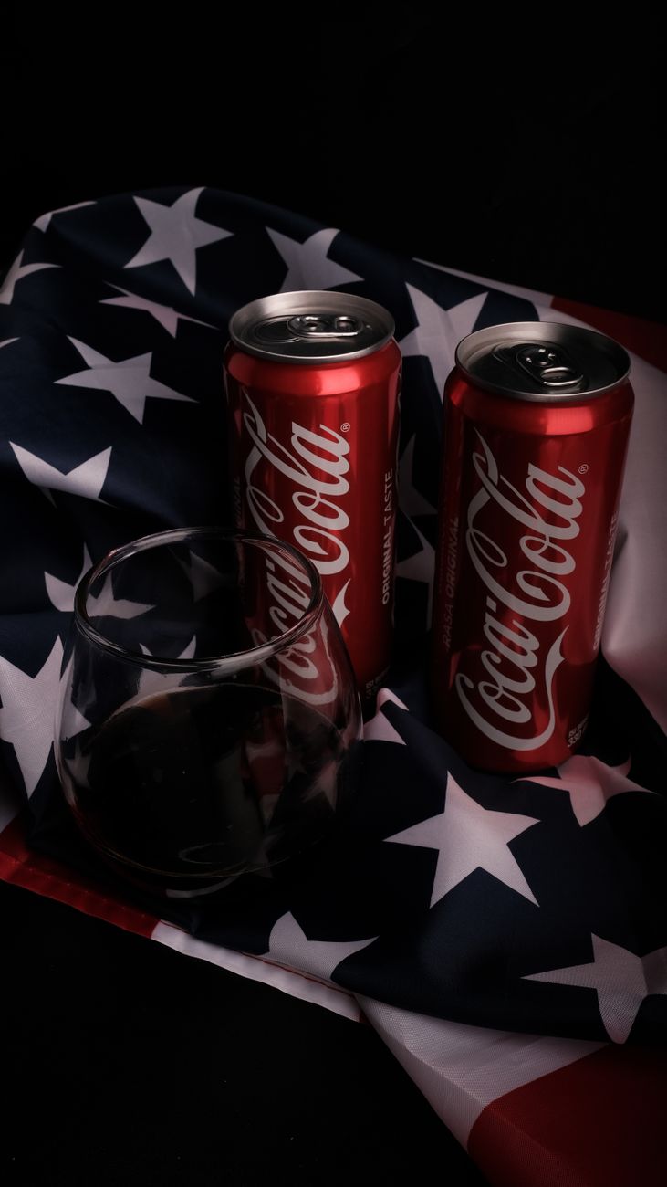 Coca-Cola cans and drink glass on an American flag backdrop in a close-up shot.