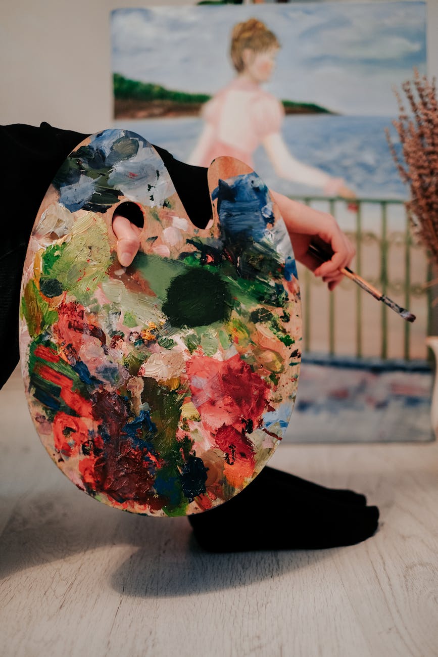 A close-up of an artist's hand holding a vibrant paint palette indoors, with a painting in the background.