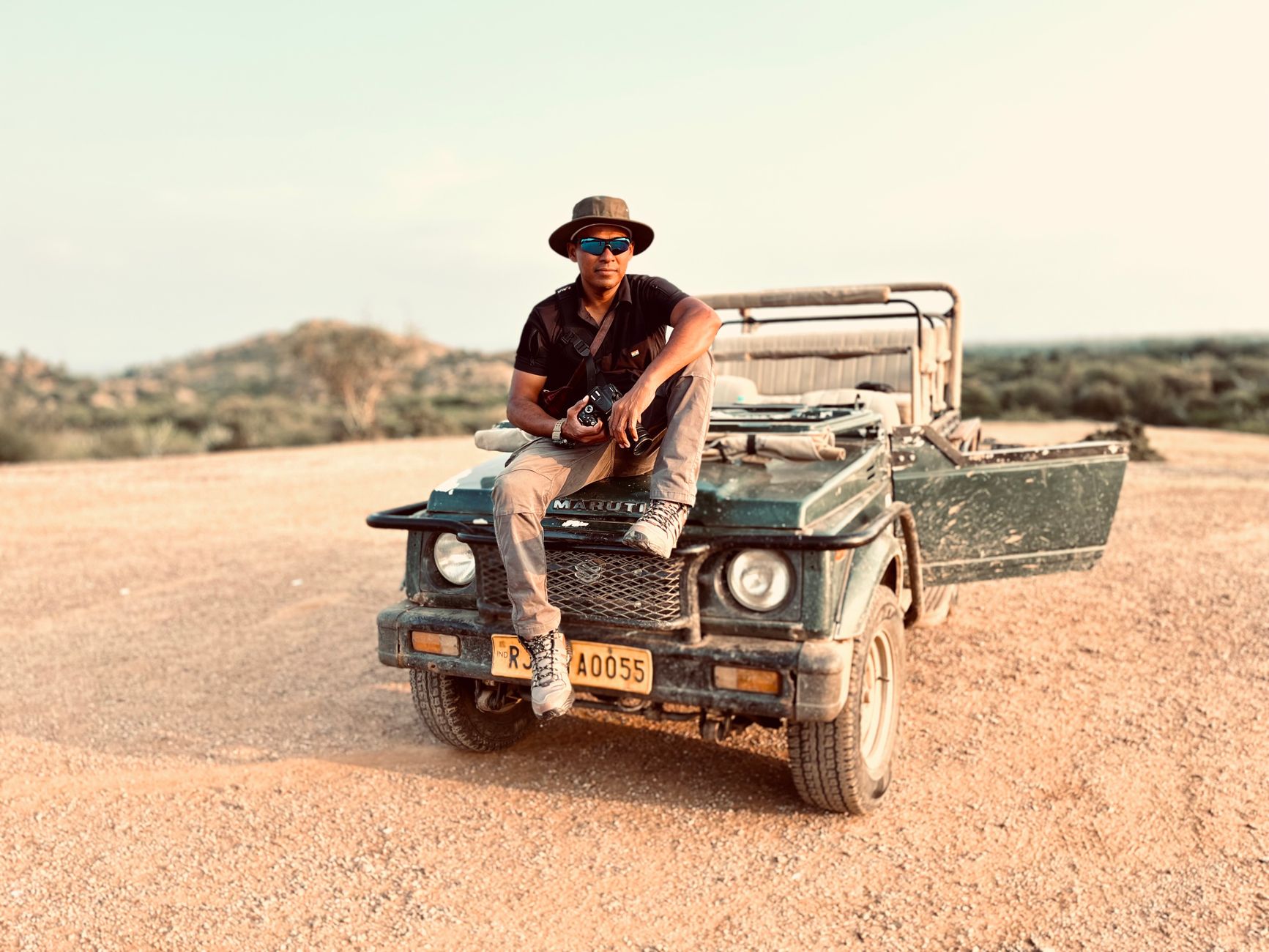 A man in sunglasses sits on a jeep in a dry, rugged landscape, exuding adventure.