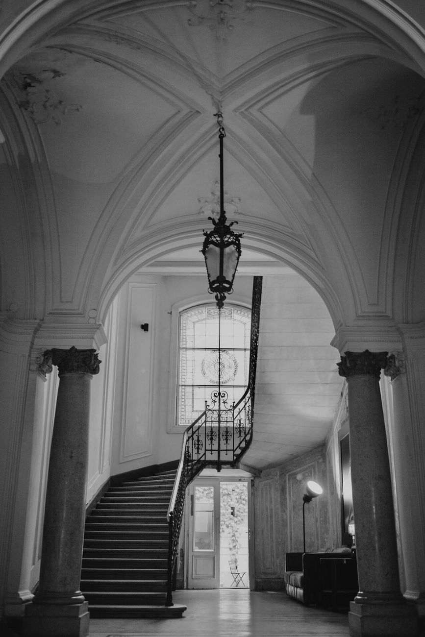 A black and white photo of a classic staircase inside a historic building, featuring arches and a chandelier.