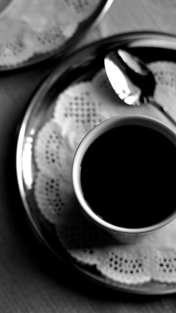 A stylish black and white image of a coffee cup on a doily-covered plate with a spoon.