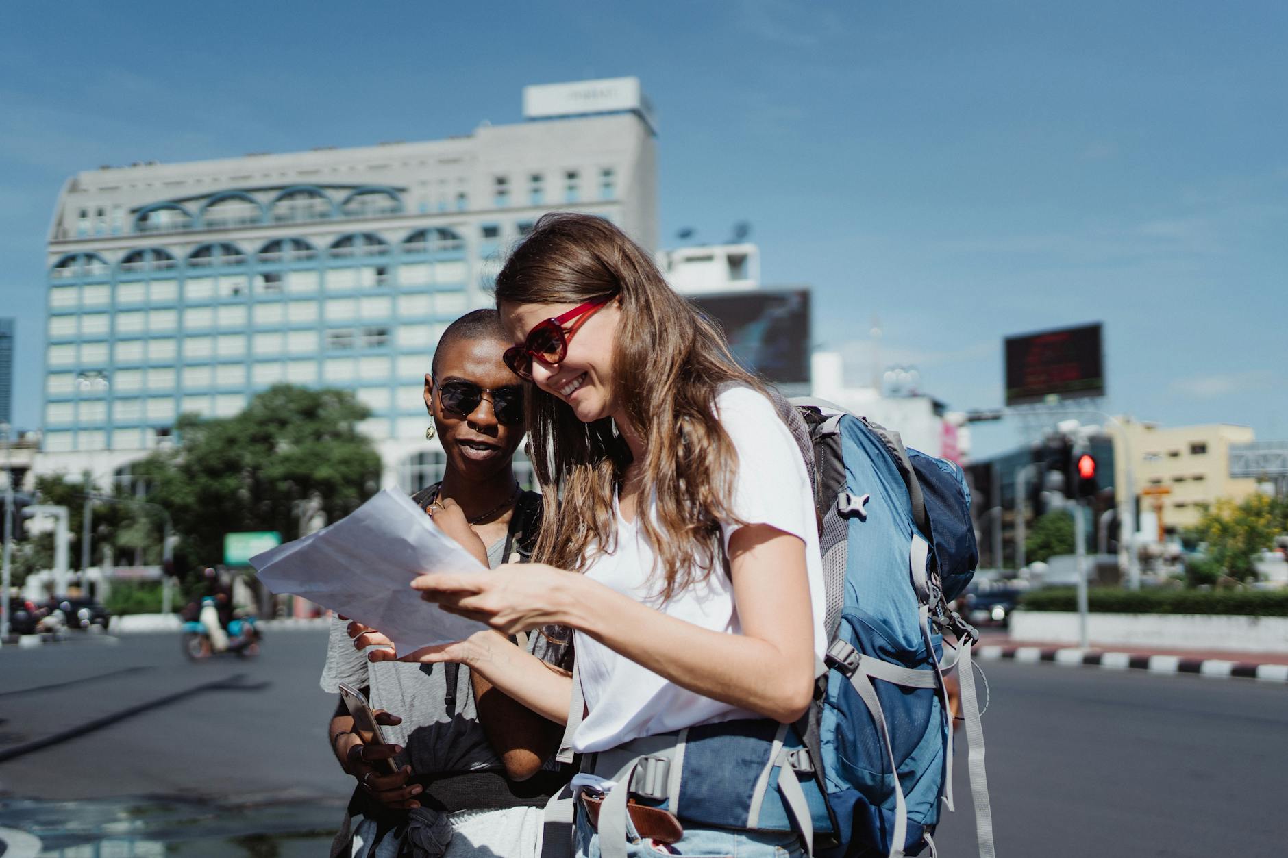 Two women tourists with backpacks check a map on a city street under a bright sky.