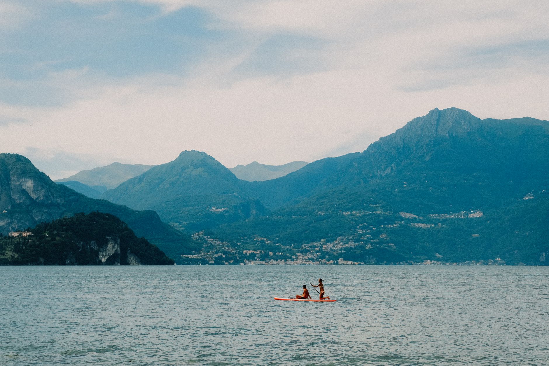 Two people kayaking on a tranquil lake surrounded by scenic mountains under a cloudy sky.