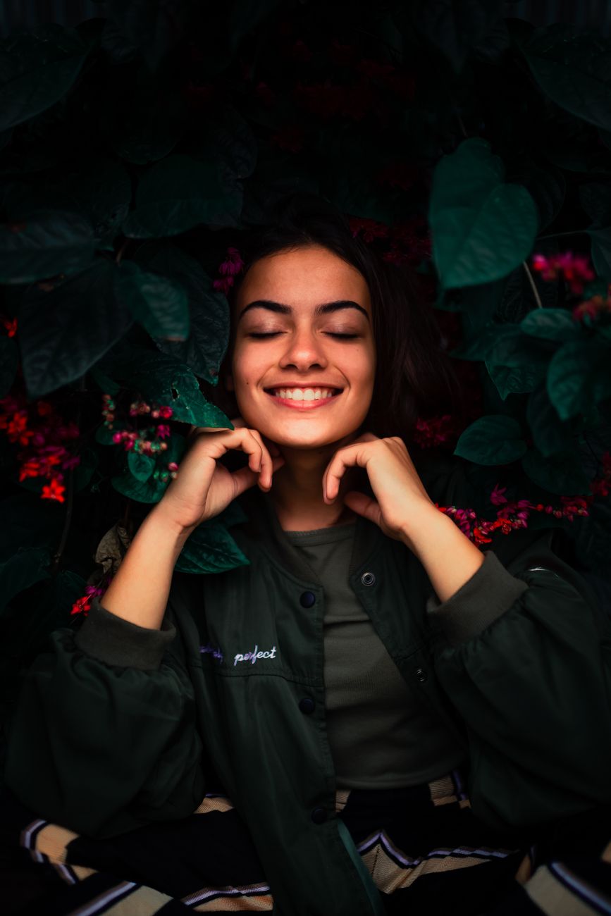An adult woman with eyes closed smiling amidst lush greenery and vibrant flowers in an outdoor portrait.