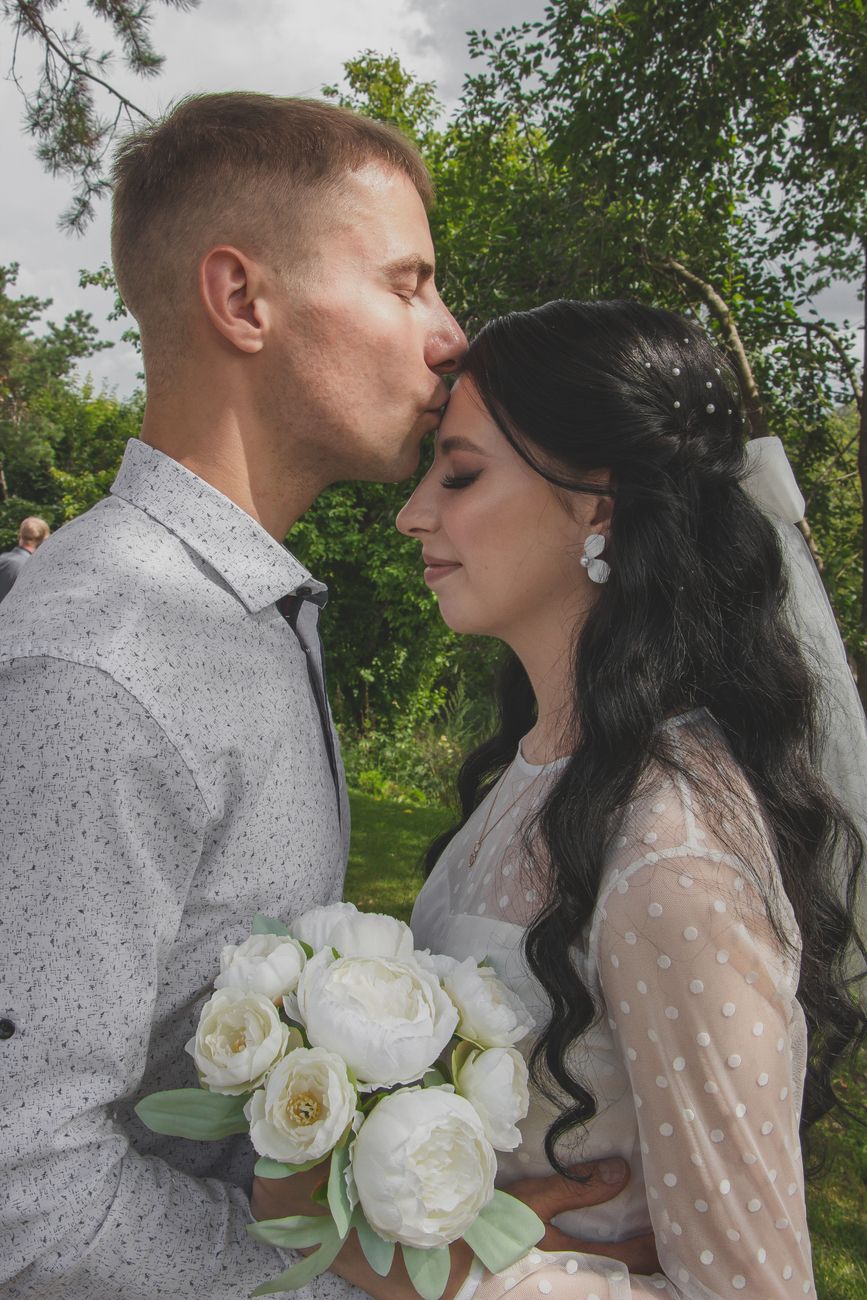 A groom kisses his bride's forehead during their outdoor wedding ceremony.