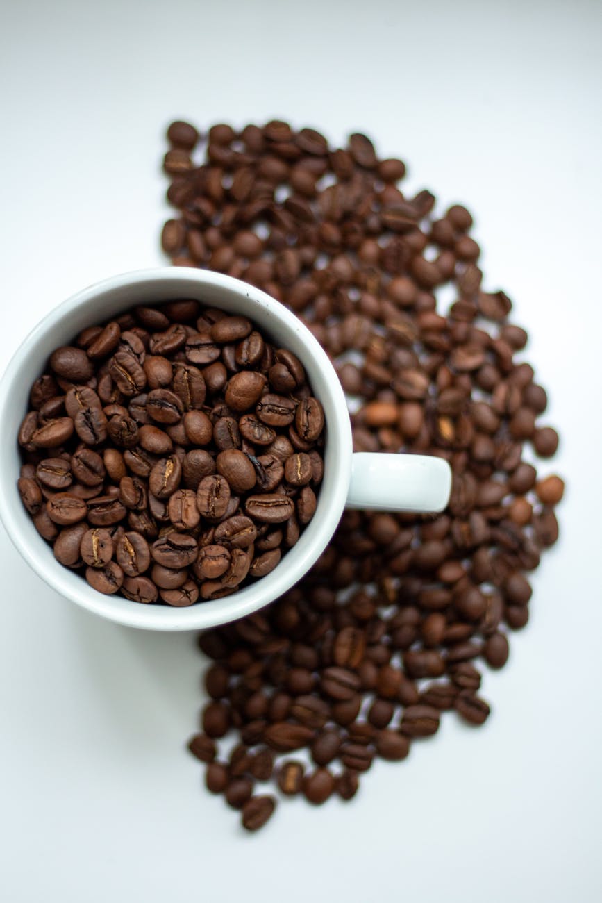 High-angle view of coffee beans spilling from a white cup on a white background, creating a rich texture.