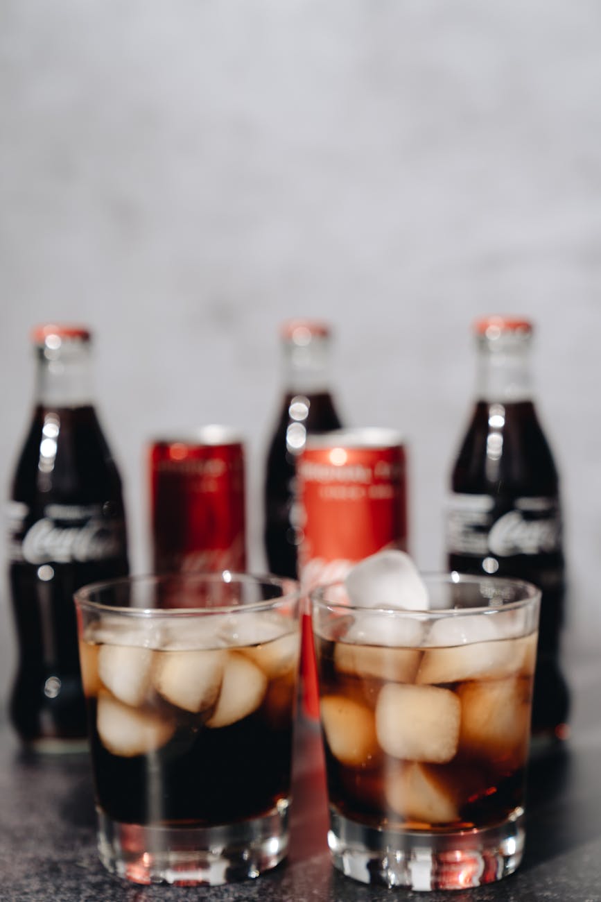 Two glasses of soda with ice cubes, featuring bottles and cans in a blurred background.